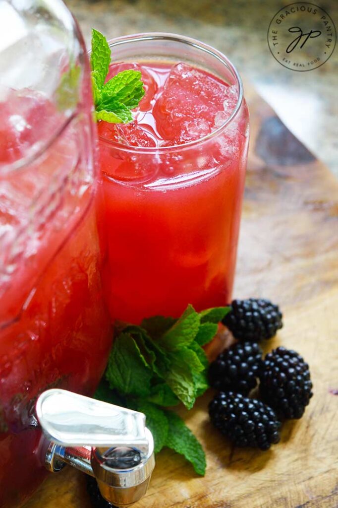 A glass of Blackberry Lemonade is garnished with fresh mint leaves and sits next to a large, glass jug of more lemonade.