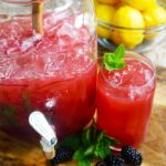 A just and glass sit on a wood cutting board, both holding Blackberry Lemonade.