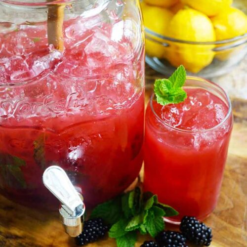 A just and glass sit on a wood cutting board, both holding Blackberry Lemonade.