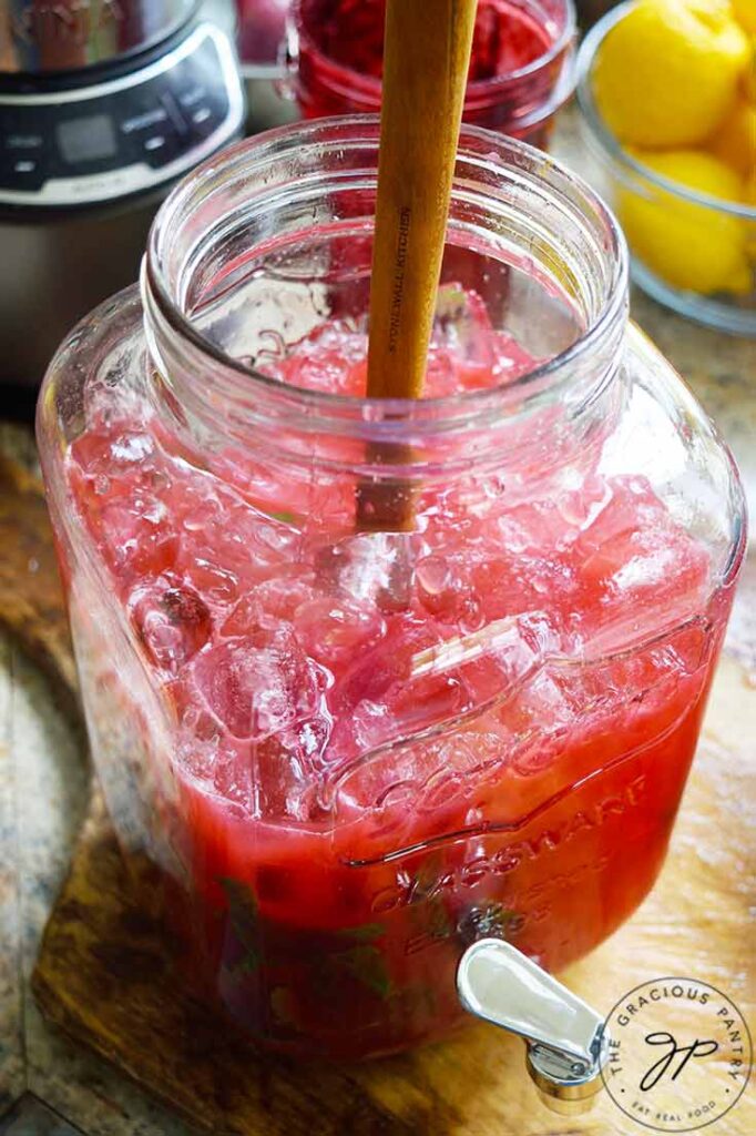 A glass beverage jug with a spigot, halfway filled with ice and Blackberry Lemonade.