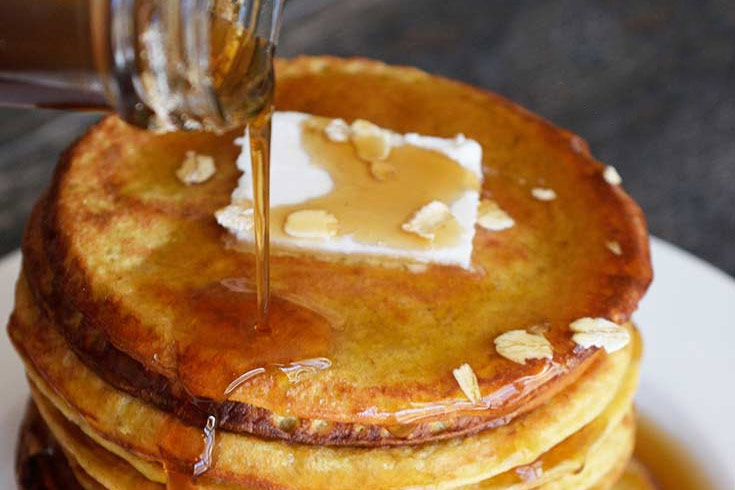 A bottle of syrup pouring over a stack of pancakes with a pat of butter on top.