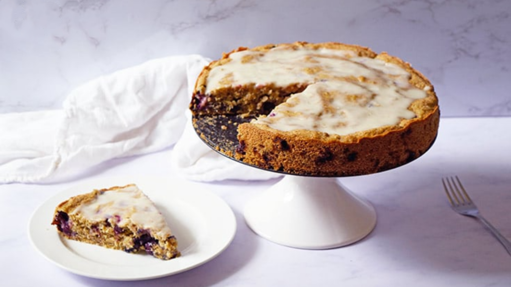 A blueberry cake sits on a cake stand with one piece removed. That slice sits a white plate next to the cake stand.