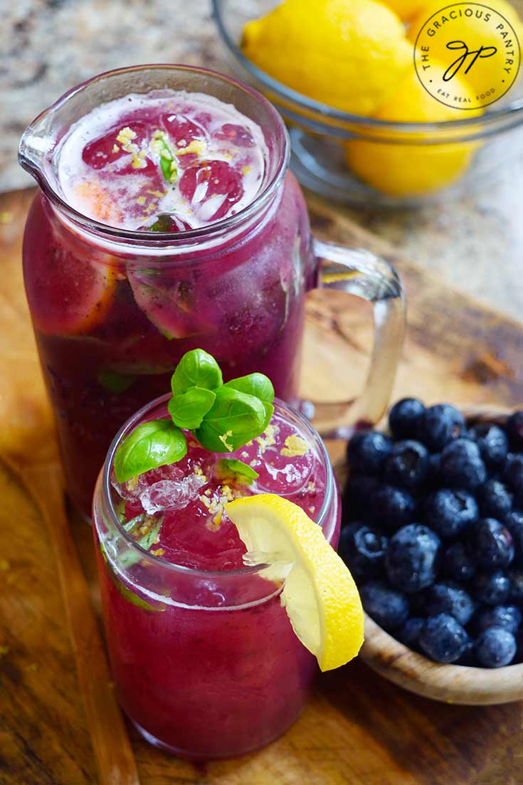 A glass pitcher and glass sit filled with Blueberry Lemonade on a cutting board. A bowl of blueberries sits to the right.