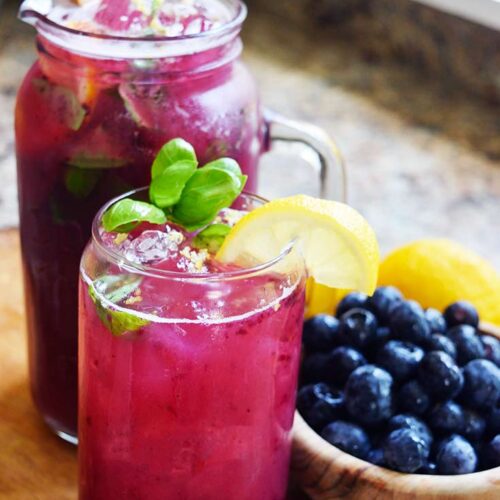 A side view of a glass and glass pitcher filled with Blueberry Lemonade sitting next to a bowl of blueberries.