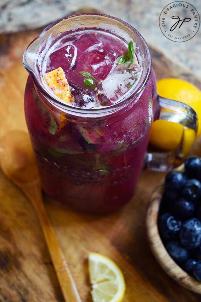 A glass pitcher full of ice and Blueberry Lemonade sits next to a wooden bowl of blueberries on a wood cutting board.
