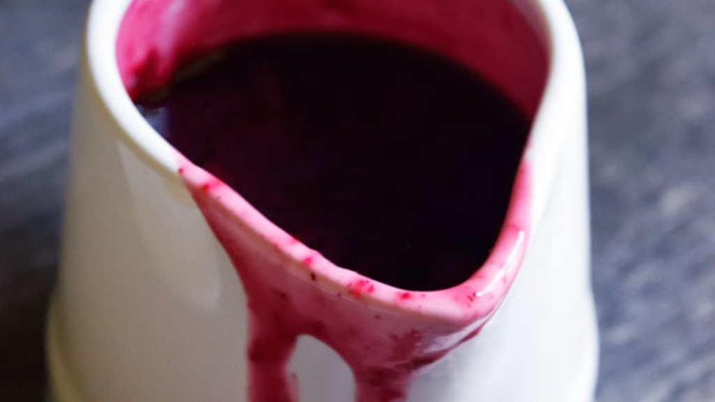 A closeup of a small white pitcher filled with blueberry syrup. Some syrup runs down the front of the pitcher.