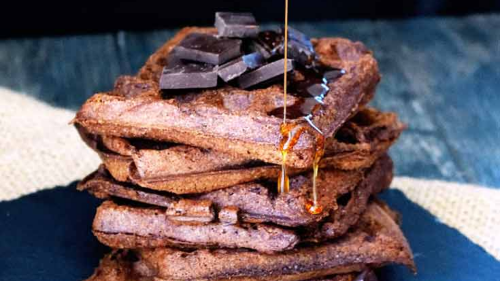 Pouring syrup over a stack of brownie waffles that are garnished with square chocolate pieces.