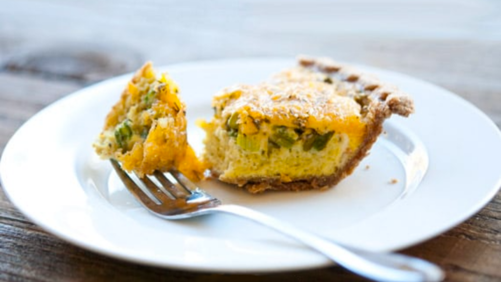A piece of cheddar asparagus quiche on a white plate. A fork has removed the tip of the triangular piece.