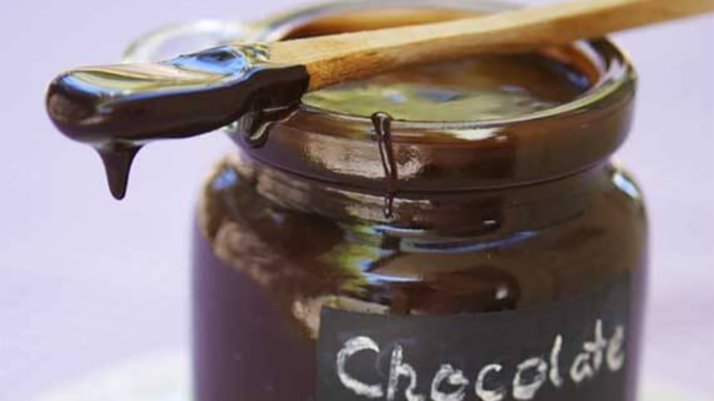 A wood spoon lays across of jar of chocolate syrup. Syrup drips from the end of the spoon.