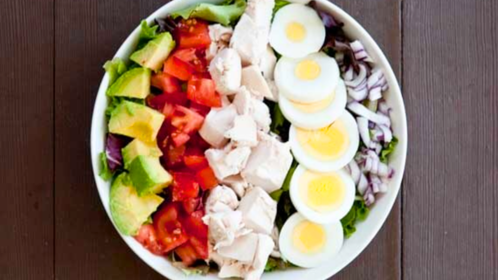 An overhead view of a cob salad in a white bowl on a wood surface.