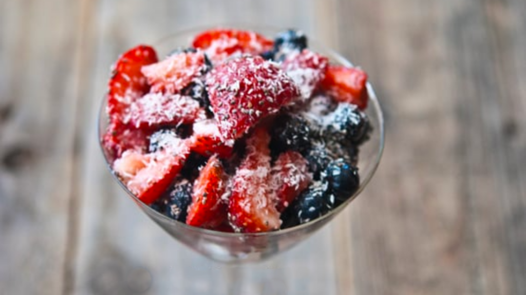 A glass bowl of coconut berry salad on a wooden background.