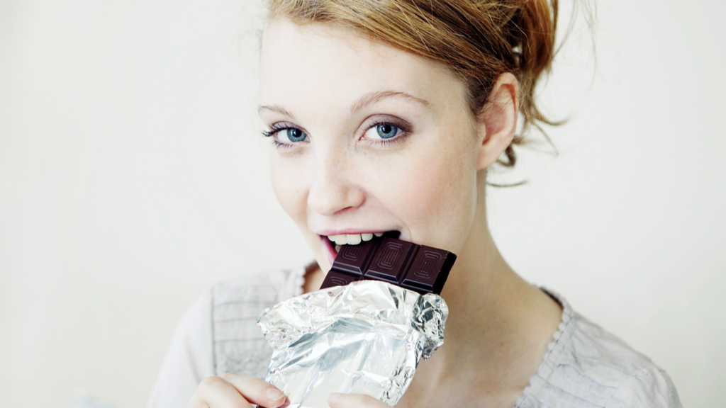 A woman takes a bite of a dark chocolate bar.