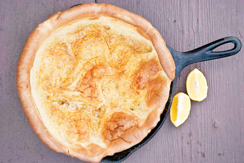 An overhead view looking down into a cast iron pan with a dutch baby pancake in it. Two lemon wedges sit to the right of the skillet.