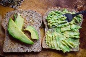 Two slices of bread lay on a wooden surface. One has avocado slices and the other is getting avocado mashed into it with a fork.