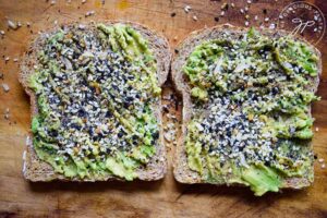 Two slices of Everything Bagel Avocado Toast lay on a wooden surface covered in seasoning.