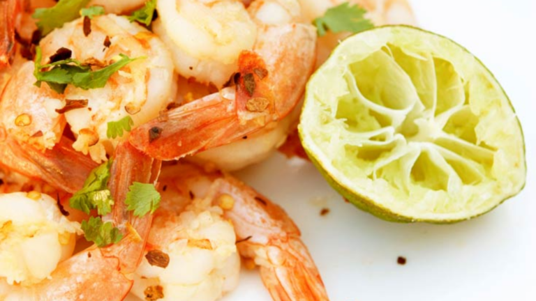 A closeup of garlic lime shrimp on a white surface with a sued lime half sitting to the side.