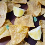 Homemade Corn Chips scattered on a cutting board with lemon wedges and chopped greens.