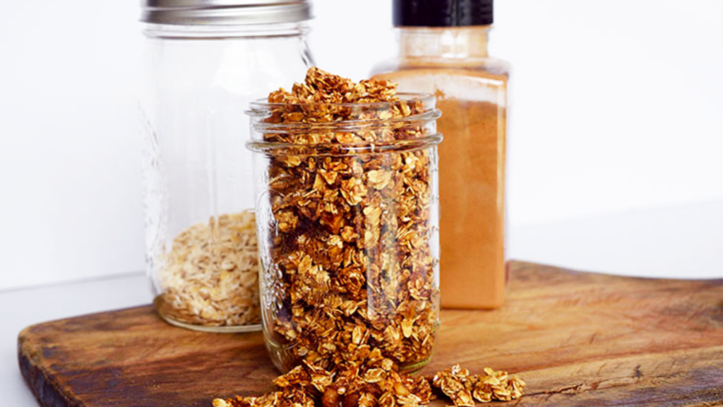 A mason jar full of homemade granola sits on a cutting board. A jar or plain oats and a bottle of cinnamon stand behind it.
