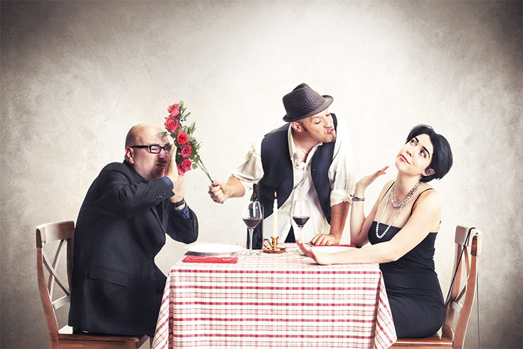 A man with flowers, shoving them in another man's face while he talks to the woman at the table.