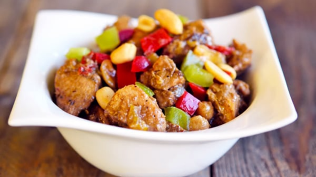 A white bowl holds a serving of kung pao chicken and is garnished with peanuts.