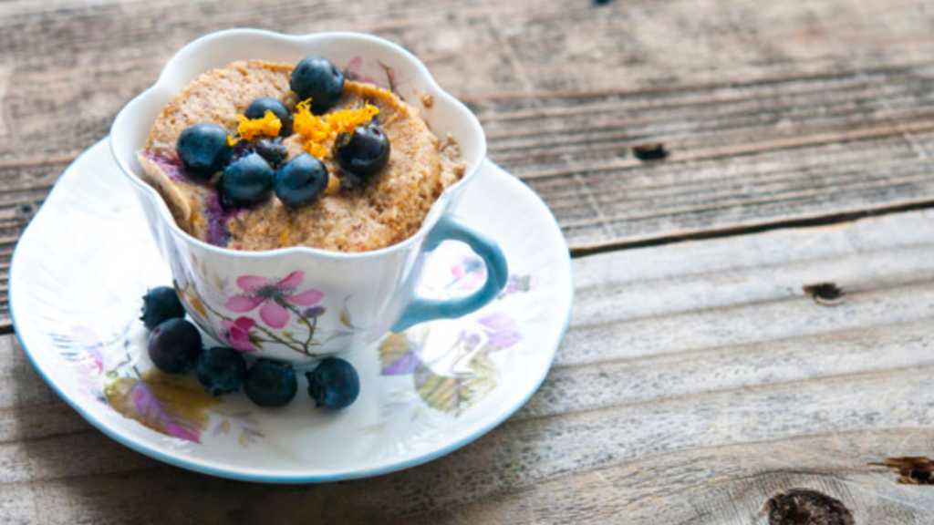 A lemon blueberry mug cake in a teacup with a saucer is garnished with fresh blueberries and fresh lemon zest.