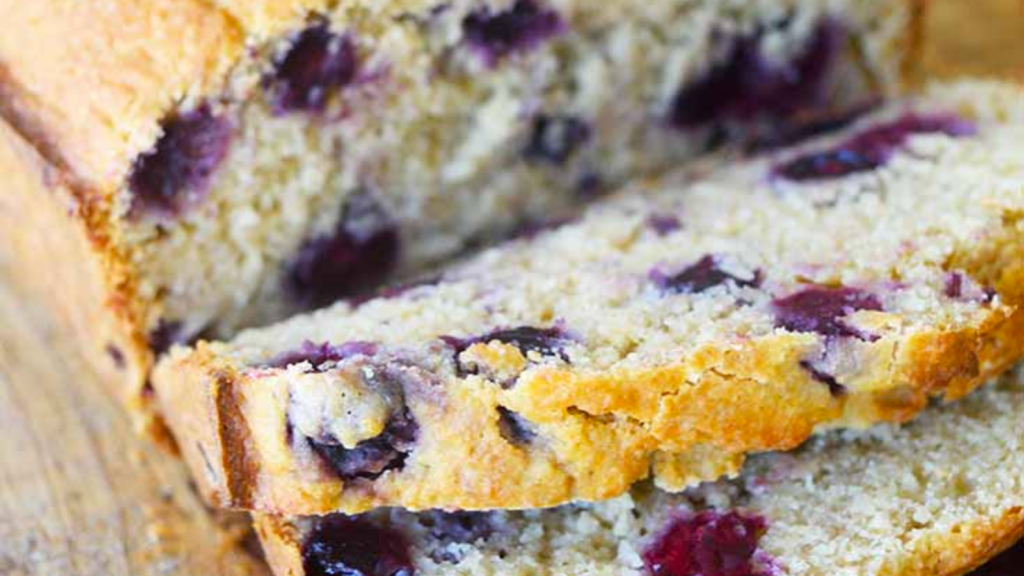 Two slices cut off of the end of a loaf of lemon blueberry bread lay on a cutting board in front of the loaf of bread.