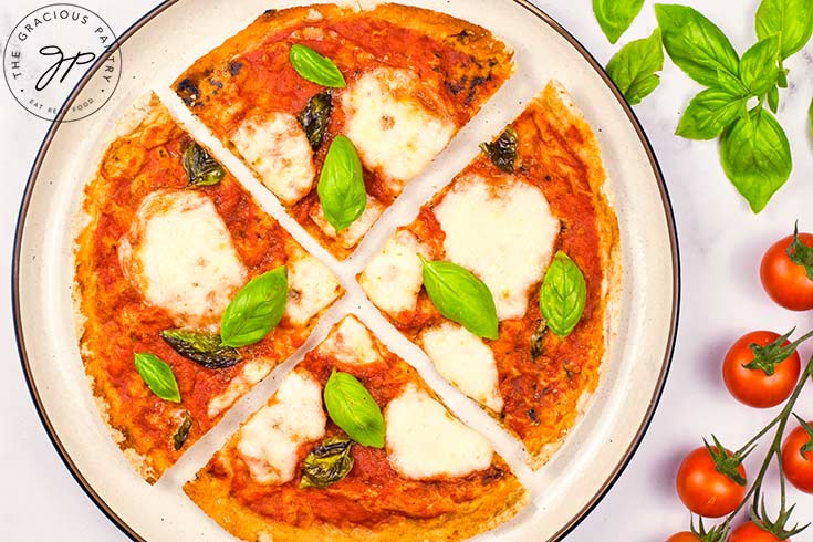 A sliced Margherita Pizza on a plate. Fresh basil and cherry tomatoes on the stem lay to the right of the plate.