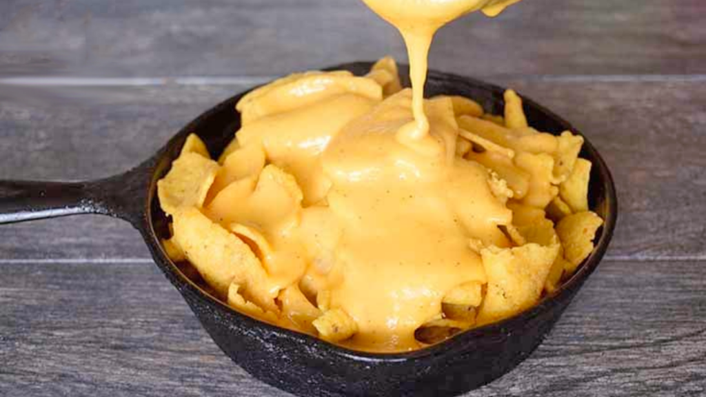 Nacho cheese sauce being poured over corn chips in a cast iron skillet.