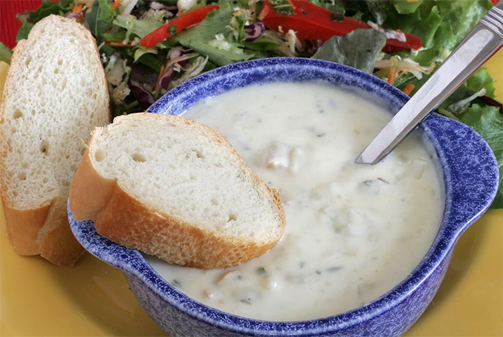A blue crock filled with New England clam chowder. Two pieces of bread rest to the side.