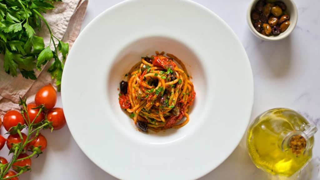 An overhead view of a white pasta bowl with a wide rim. In the center sits a next of pasta puttanesca.