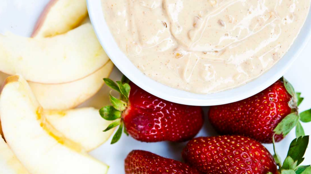 A white bowl of peanut butter fruit dip surrounded by apple slices and strawberries.