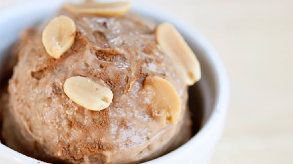 A closeup of a scoop of peanut butter n'ice cream in a small white bowl. It's garnished with fresh peanuts.