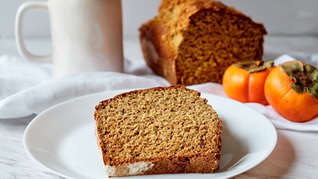 A slice of persimmon bread sits on a white plate with the persimmon loaf sitting behind it.