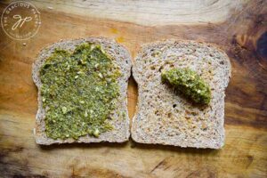 Two slices of whole-grain bread on a cutting board. One has pesto sauce spread over it, the other has a dollop of pesto sauce resting on it.