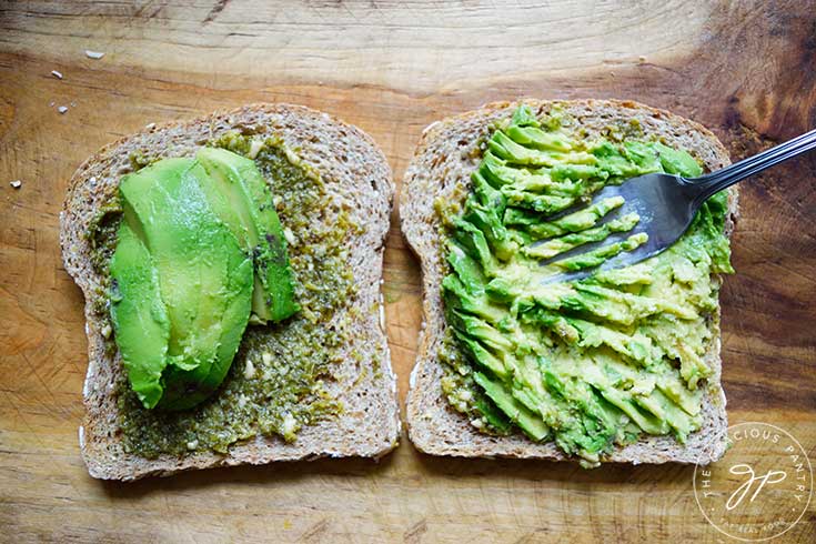 Avocado slices being mashed into slices of bread with pesto sauce on them.