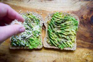 Sprinkling parmesan cheese over the top of two slices of avocado toast.