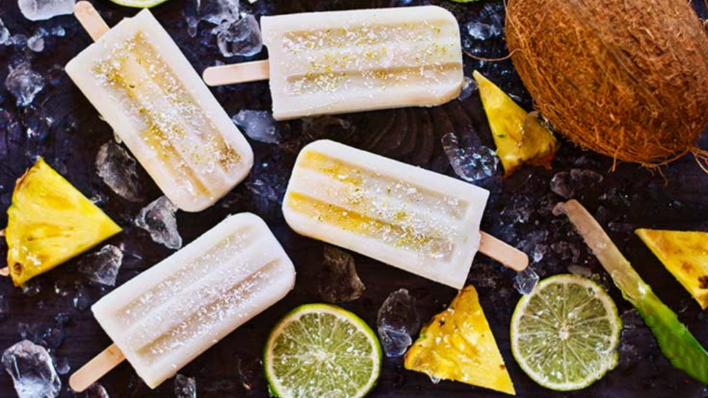 A dark gray background holds four pineapple popsicles, ice cubes, lime slices, pineapple wedges and a coconut, all artfully arranged.