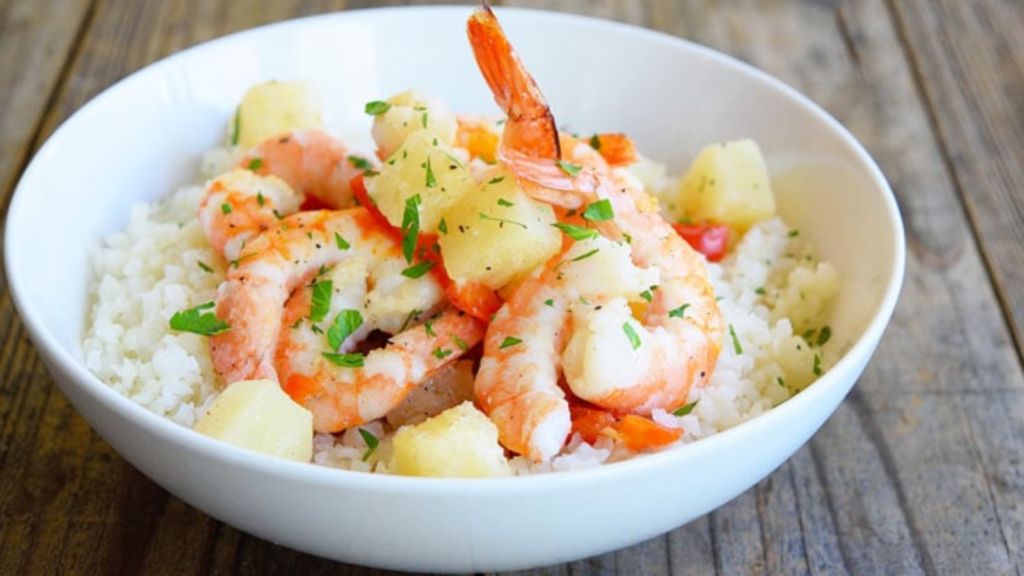 A white bowl holds sheet pan pineapple shrimp garnished with fresh greens.