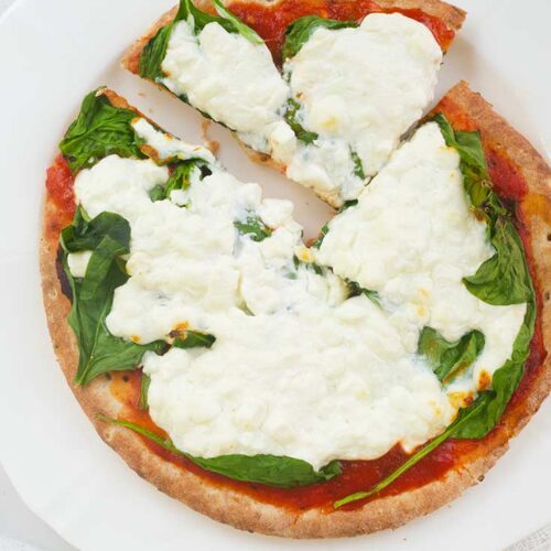 A single slice of Pita Pizza sits pulled back from the rest of the pizza on a white plate and background.