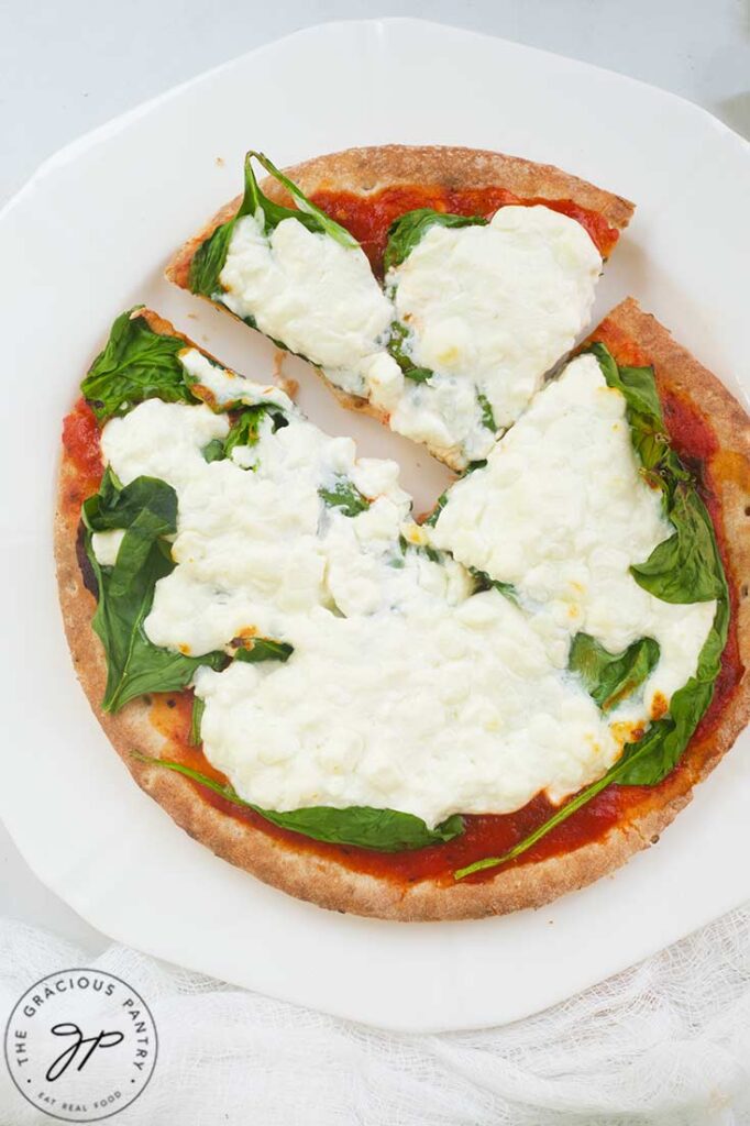 A single slice of Pita Pizza sits pulled back from the rest of the pizza on a white plate and background.