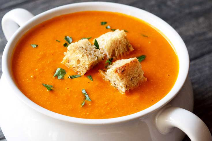 A white crock filled with Instant Pot tomato soup, garnished with three bread crumbs and fresh herbs.