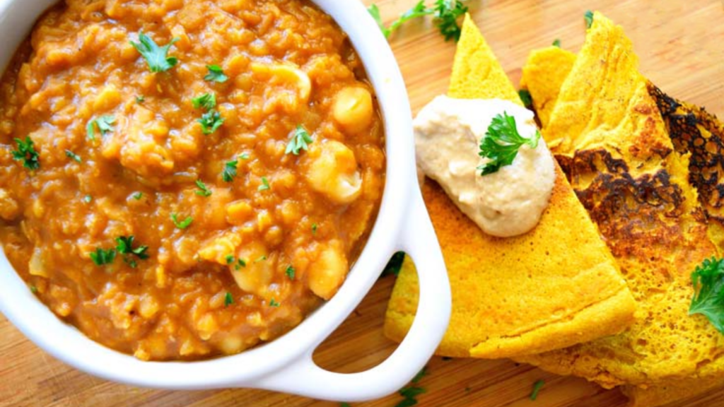 A white crock is filled with red lentil stew. Cut socca flatbread pieces lay to the right of the bowl.