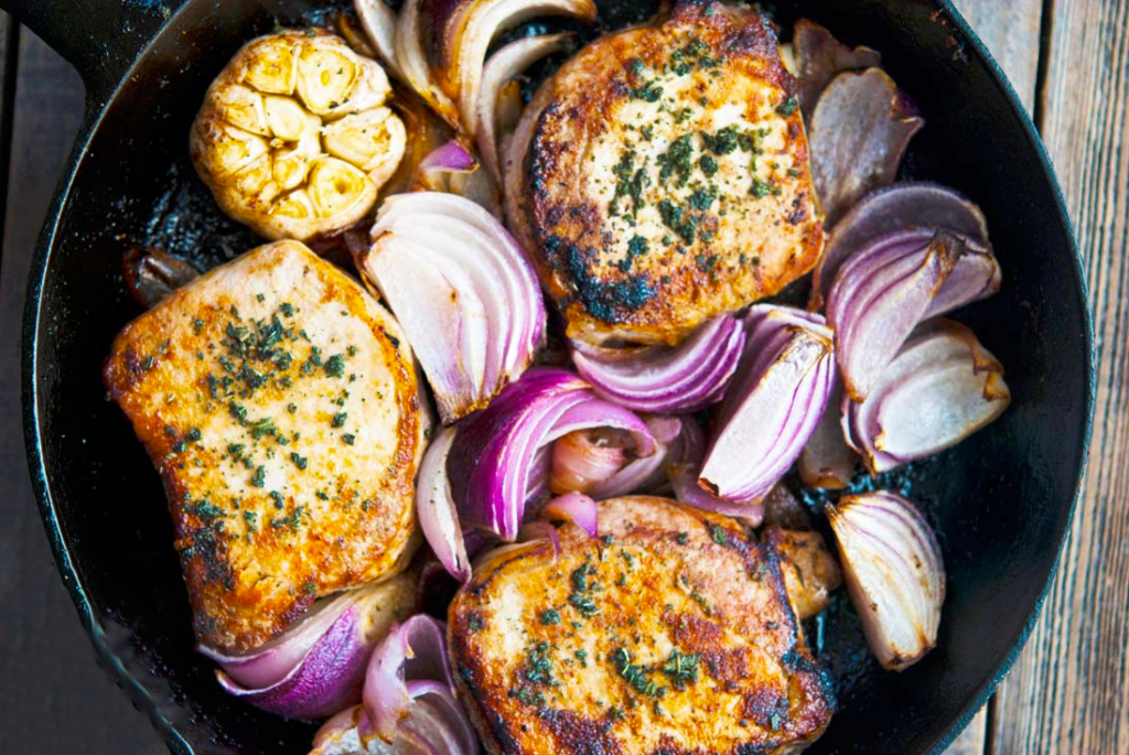 An overhead view of a cast iron skillet filled with three browned pork chops, chunks of red onion and a whole head of garlic.