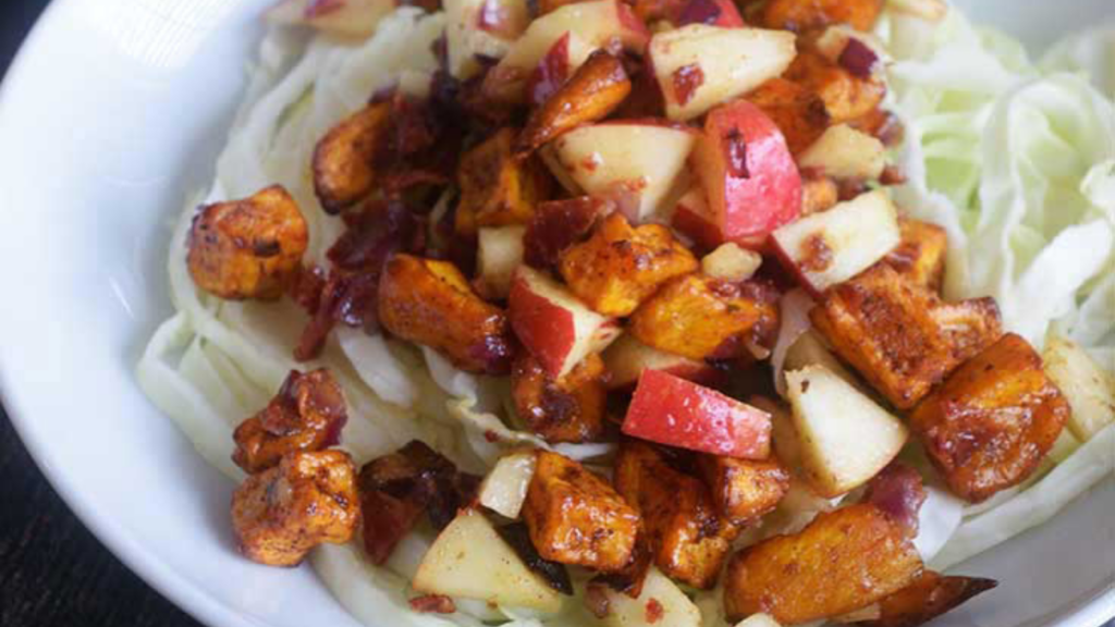 A white bowl holds a serving of roasted sweet potato salad on a bed of shredded cabbage.