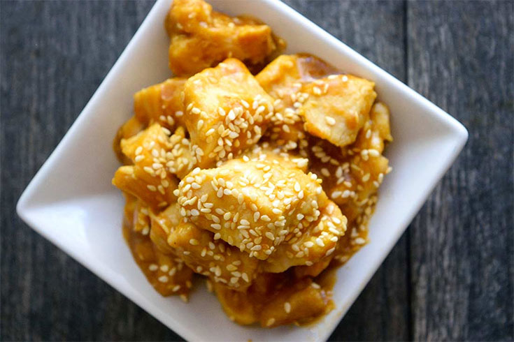 An overhead view of a square, ceramic, white dish holding a serving of sesame chicken garnished with sesame seeds.