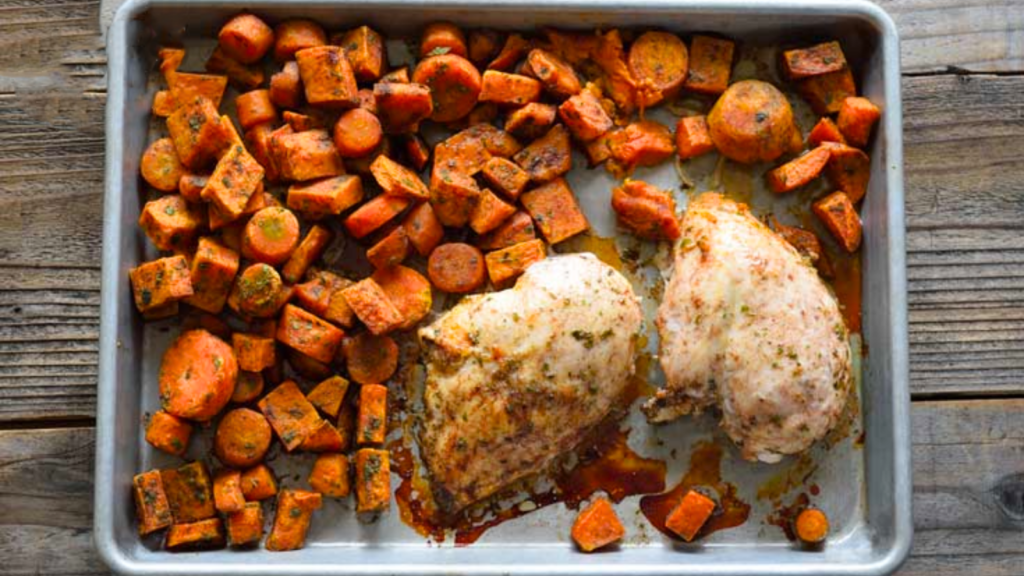 An overhead view of a sheet pan holding two cooked chicken breasts and roasted, chopped carrots.
