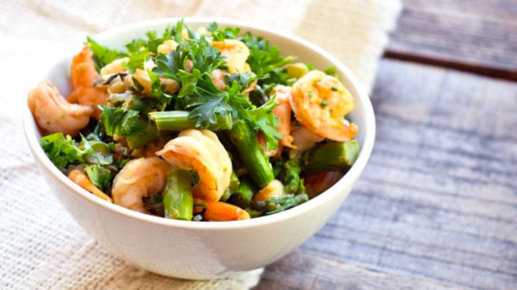A white bowl is filled with this shrimp asparagus skillet recipe. It shows shrimp, asparagus pieces and some fresh greens.