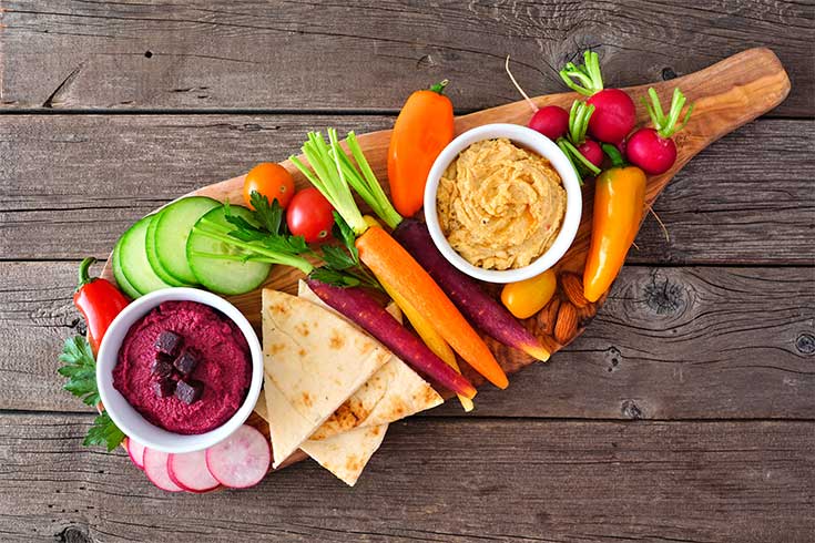 An overhead view of a charcuterie board with veggies, pita chips, and two bowls of dip.