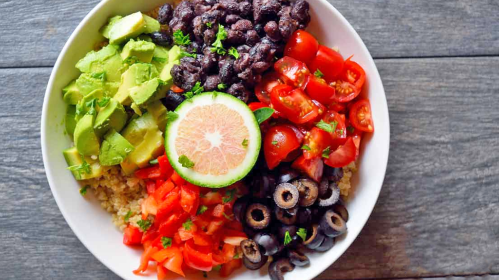An overhead view of a white bowl filled with quinoa, avocado chunks, black beans, tomatoes, olives and red peppers.