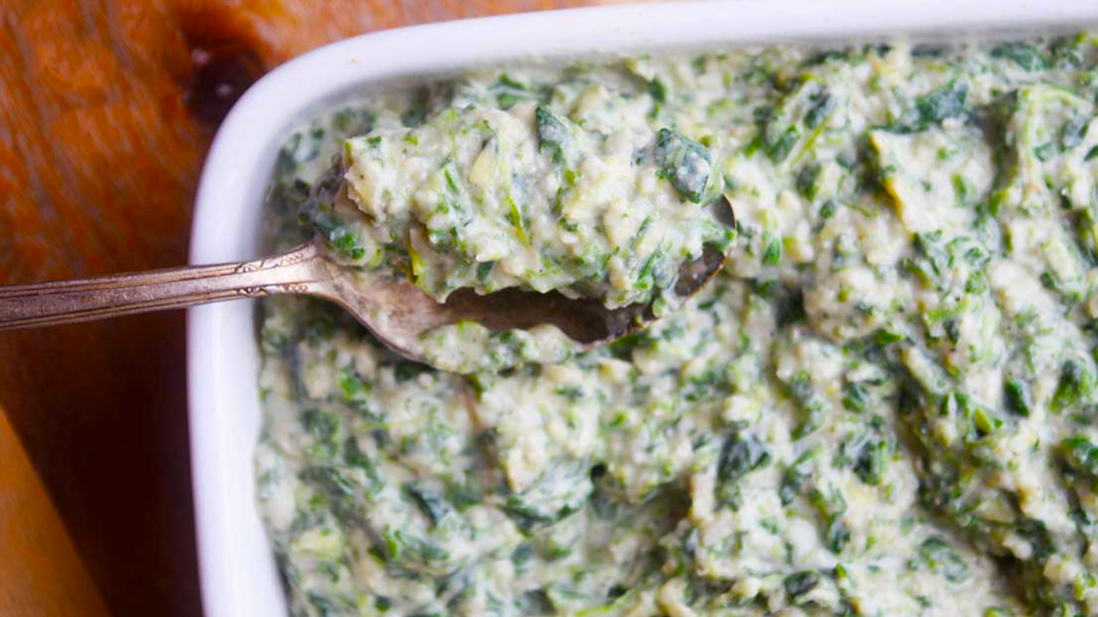 A spoon lifts a bit of spinach artichoke dip out of a white dish.
